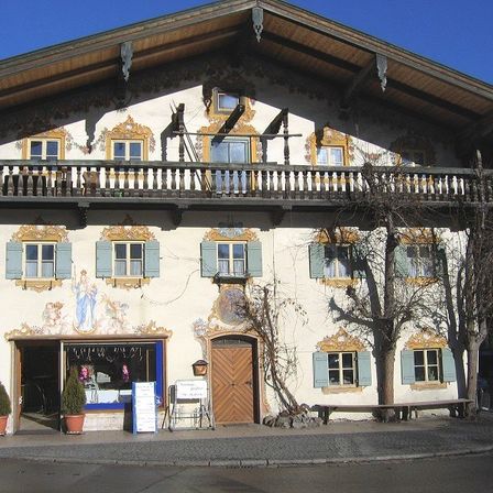 Ein mehrstöckiges Gebäude mit aufwendigen Verzierungen und einer Holztür hat mehrere Fenster mit blauen Jalousien. Davor stehen zwei Topfpflanzen.