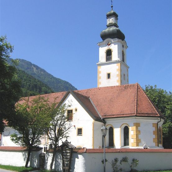 Eine weiße Kirche mit einem braunen Dach und einem hohen Turm steht vor einem Berg. Ein Uhr schmückt den Turm. Bäume flankieren die Kirche, und eine Straßenlaterne steht davor.