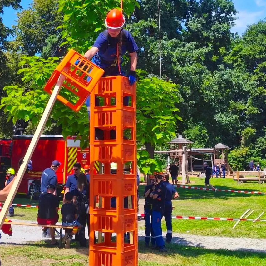 Eine Person in einem Gurt klettert auf einen Turm aus orangefarbenen Kisten in einem Park mit Zuschauern und einem Feuerwehrauto im Hintergrund.