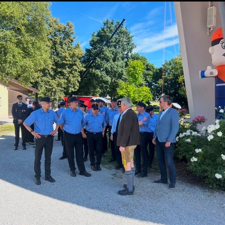 Eine Gruppe von Feuerwehrleuten in blauen Uniformen steht vor einem Gebäude mit einer großen Figur an der Seite. Ein Mann in einem Anzug spricht mit ihnen.