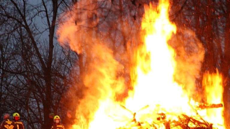 Ein großes Lagerfeuer brennt in einem bewaldeten Gebiet, wobei Feuerwehrleute in der Nähe stehen. Das Feuer ist von Bäumen umgeben und gibt viel Rauch ab.