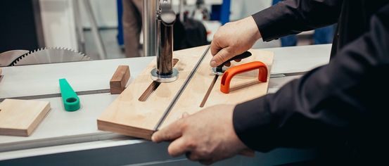 Eine Person sägt ein Holzbrett mit einer Säge, wobei ein Metallständer verwendet wird. Eine andere Person steht dahinter und hält einen Trolley. Der Raum hat weiße Wände und einen weißen Boden.