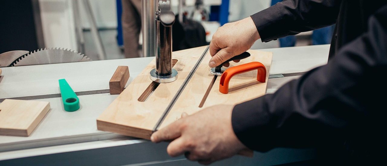 A person is cutting a wooden plank with a saw, using a metal stand. Another person is standing behind, holding a trolley. The room has white walls and a white floor.