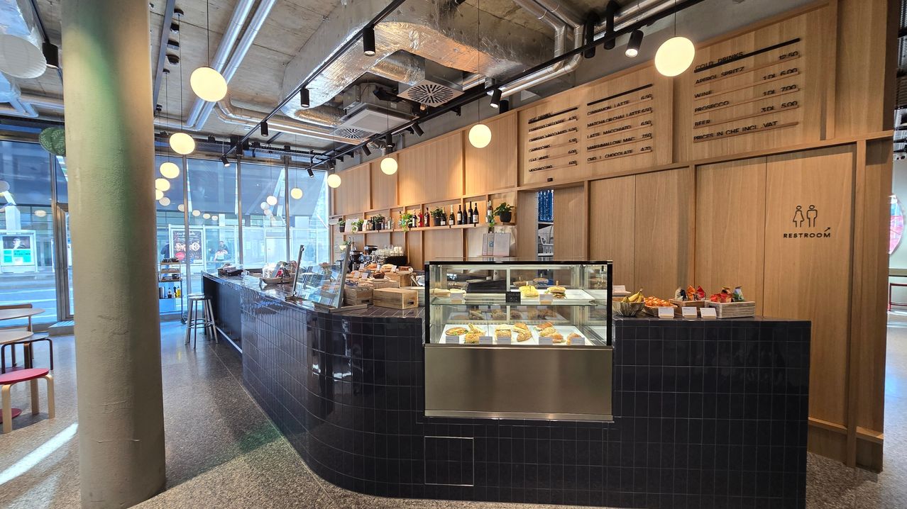 Das Innere eines Bäckereiladens mit einer gebogenen schwarzen Theke, einer Glasvitrine mit Backwaren, einem Regal mit Flaschen und einer Menütafel.