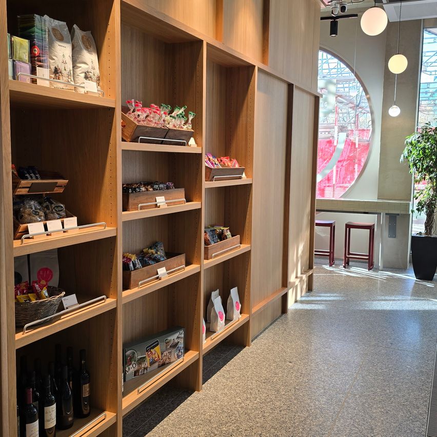 Eine Innenausstellung verschiedener Lebensmittel auf Holzregalen. Ein rotes Fenster und ein Topfpflanzen stehen auf der rechten Seite. Zwei Hocker stehen in der rechten Ecke.