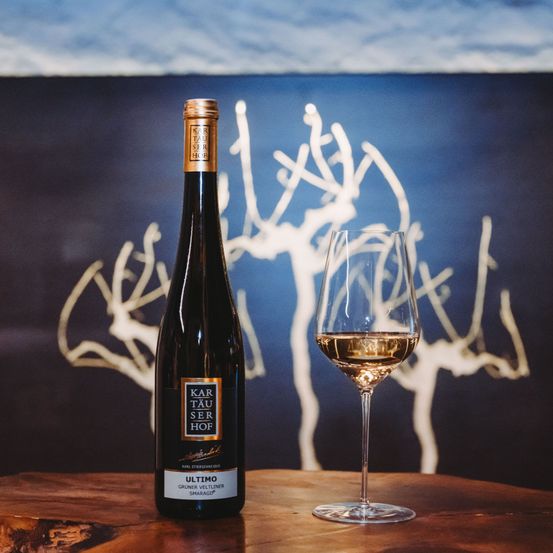 Eine Flasche Karl Tauser Hof's ULTIMO Wein steht neben einem Glas Weißwein. Im Hintergrund befindet sich abstrakte Kunst von Bäumen.