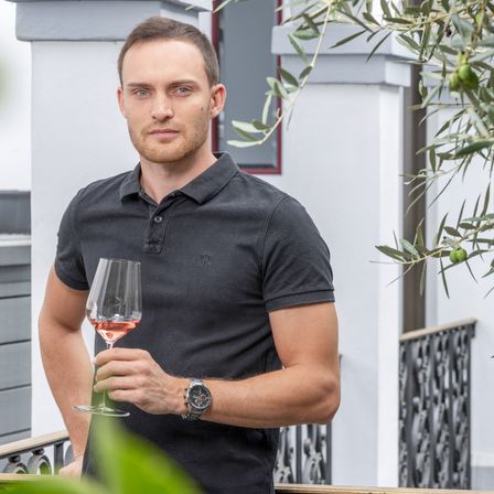 Ein Mann mit einer Uhr hält ein Glas Roséwein auf einem Balkon mit einem Olivenbaum.
