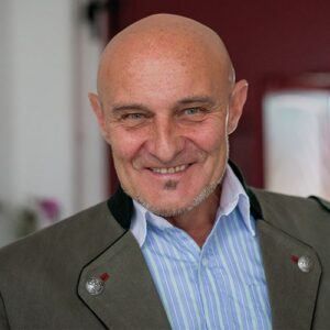 Ein Mann mit kahlem Kopf und Bart lächelt in die Kamera. Er trägt ein blau-weiß gestreiftes Hemd unter einer grauen Jacke mit rotem Kragen. Er steht vor einer roten Tür.