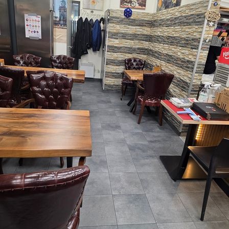 Ein leeres Restaurant mit Holztischen und Stühlen. Es gibt Lederstühle, einen Kühlschrank und eine Kleiderstange. Die Wände sind aus Ziegelsteinen, und es hängen Poster daran. Es gibt einen schwarzen Tisch mit Büchern und einem Handy darauf.