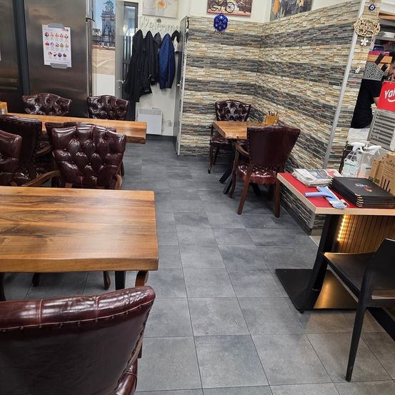 Ein leeres Restaurant mit Holztischen und Stühlen. Es gibt Lederstühle, einen Kühlschrank und eine Kleiderstange. Die Wände sind aus Ziegelsteinen, und es hängen Poster daran. Es gibt einen schwarzen Tisch mit Büchern und einem Handy darauf.