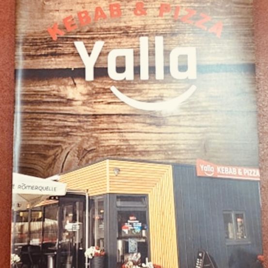 Ein Poster für ein Restaurant namens Yalla, mit einem hölzernen Hintergrund und einem lächelnden Logo. Es zeigt ein kleines Lokal mit gelber Fassade, einem Vordach und schwarzem Außenbereich. Draußen sind Tische, Stühle und Blumen zu sehen.