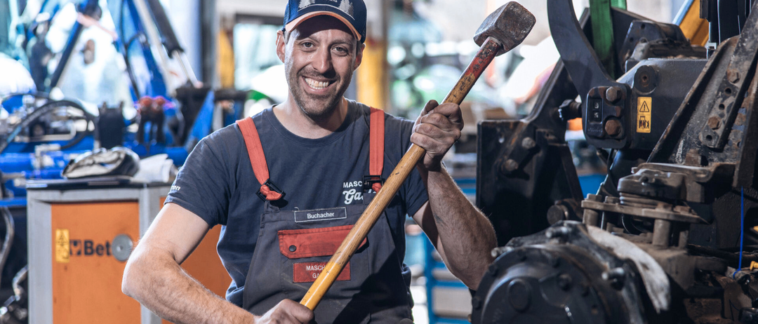 Ein lächelnder Mechaniker mit einer Husqvarna-Kappe und einem grauen Hemd mit orangefarbenen Hosenträgern hält einen großen Hammer in einer Werkstatt.
