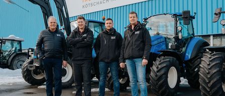Four men stand in front of blue tractors, all dressed in black jackets. Behind them is a blue building with a white banner that reads Industriepark Kötschach-Mauthen.