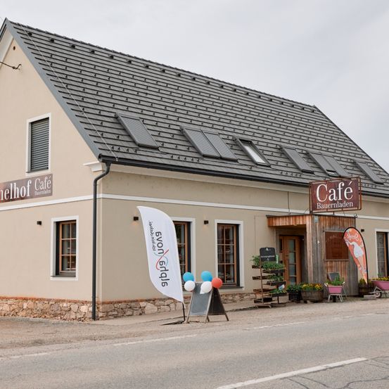 Ein Café-Gebäude mit grauem Dach und Fenstern, mit einem Café-Schild und einem Banner davor. Draußen befinden sich Luftballons und Topfpflanzen.