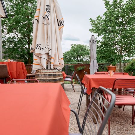 Ein Außenbereich mit mehreren Tischen und Stühlen, mit orangefarbenen Tischdecken und Sonnenschirmen in der Mitte. Bäume und ein Zaun sind im Hintergrund.