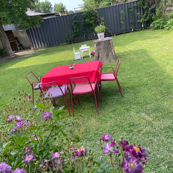Ein Garten mit einem roten Tisch und Stühlen auf dem Rasen. Es gibt einen Baumstumpf mit einem Topf. Lila Blumen und Bäume sind in der Umgebung.