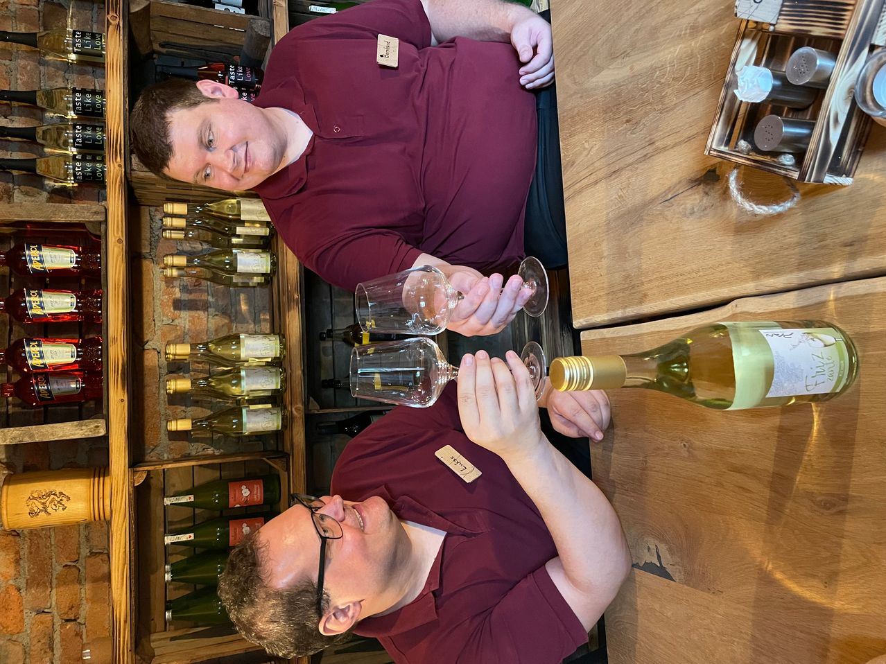 Zwei Männer sind in einem Weinladen. Ein Mann lächelt und der andere hält zwei Wein glasses. Sie gießen Wein aus einer Flasche in die Gläser. Dahinter befinden sich Regale mit vielen Weinflaschen.