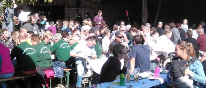 Eine große Gruppe von Menschen sitzt im Freien an Tischen. Viele tragen grüne Hemden. Eine Frau hält ein Baby, während ein Kind an einem Tisch sitzt.