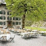 Ein großes Gebäude mit mehreren Fenstern und grünen Fensterläden ist von einem großen Baum umgeben. Das Gebäude verfügt über einen Außenbereich mit vielen Tischen und Stühlen.
