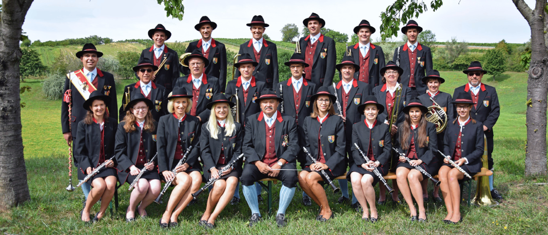 Eine Musikband mit verschiedenen Instrumenten, darunter Flöten und Trompeten, posiert für ein Gruppenfoto auf einer Grasfläche. Die Mitglieder tragen formelle Kleidung mit roten Westen und schwarzen Hüten. Bäume und Grünfläche umgeben das Gelände.