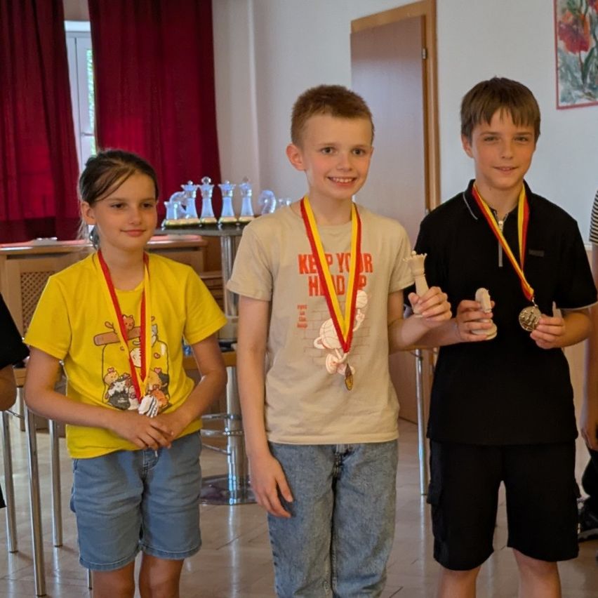 Three children wearing medals stand together. A girl in yellow, a boy in beige, and another boy in black. They are indoors, with trophies and a painting behind them.