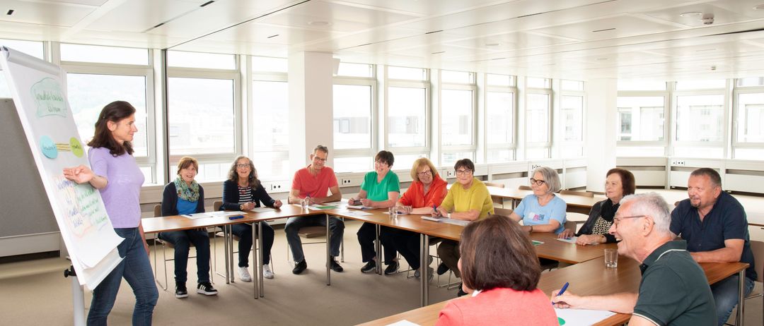 Eine Gruppe von Menschen sitzt an einem langen Tisch in einem Konferenzraum. Sie tragen Freizeitkleidung und einige tragen Brillen. Sie scheinen etwas zu diskutieren und schreiben auf Papier. Hinter ihnen befinden sich mehrere Fenster und der Raum ist gut beleuchtet.