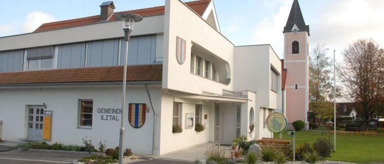 Ein modernes weißes Gebäude mit einem roten Dach und 'GEMEINDE ILTZTAL' auf der Seite geschrieben. Eine Flagge ist an der Wand montiert. Ein Turm und eine Kirche stehen dahinter.