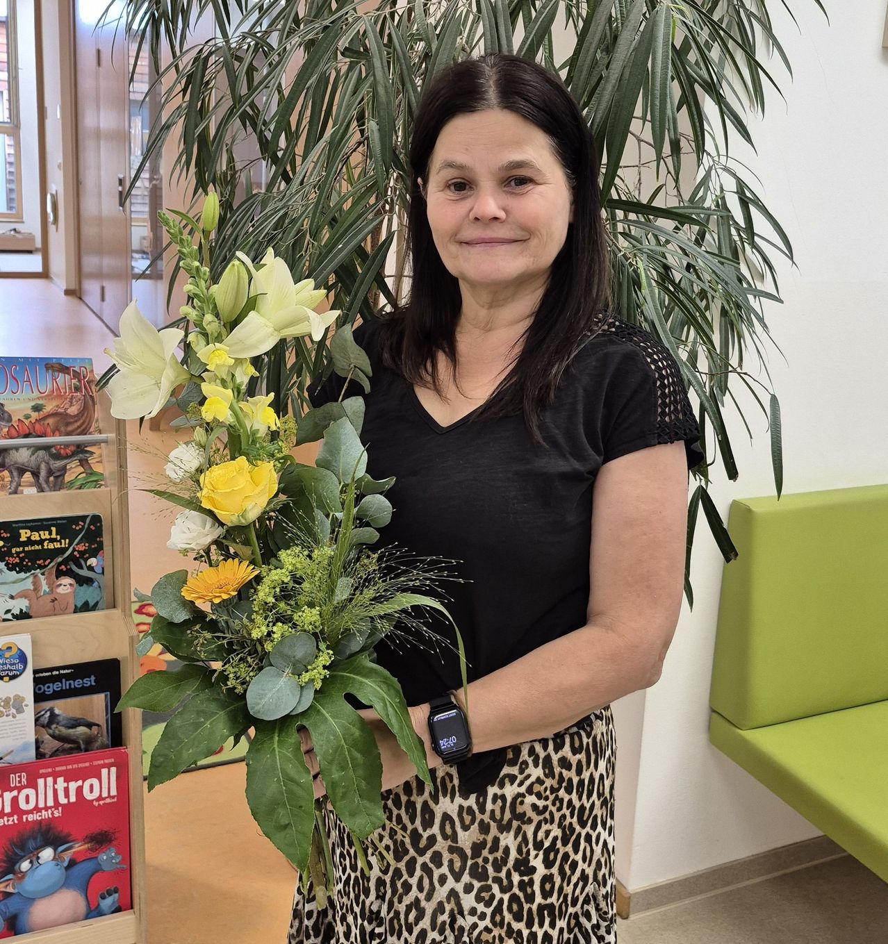 Eine Frau in einem schwarzen Shirt und einem Leopardenprintrock hält einen Blumenstrauß und lächelt in einem Raum mit Büchern und einem grünen Stuhl.