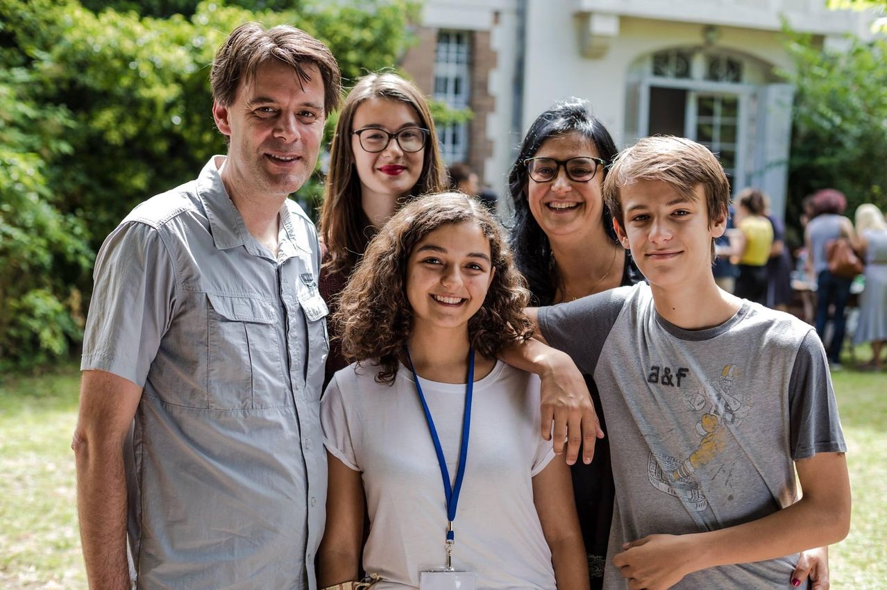 Eine Gruppe von Menschen steht vor einem Gebäude. Eine Frau in der Mitte trägt ein weißes Shirt und einen blauen Ausweis. Ein Junge rechts trägt ein graues Shirt mit einem Grafik. Dahinter sind Bäume und ein Gebäude mit Glasfenstern.