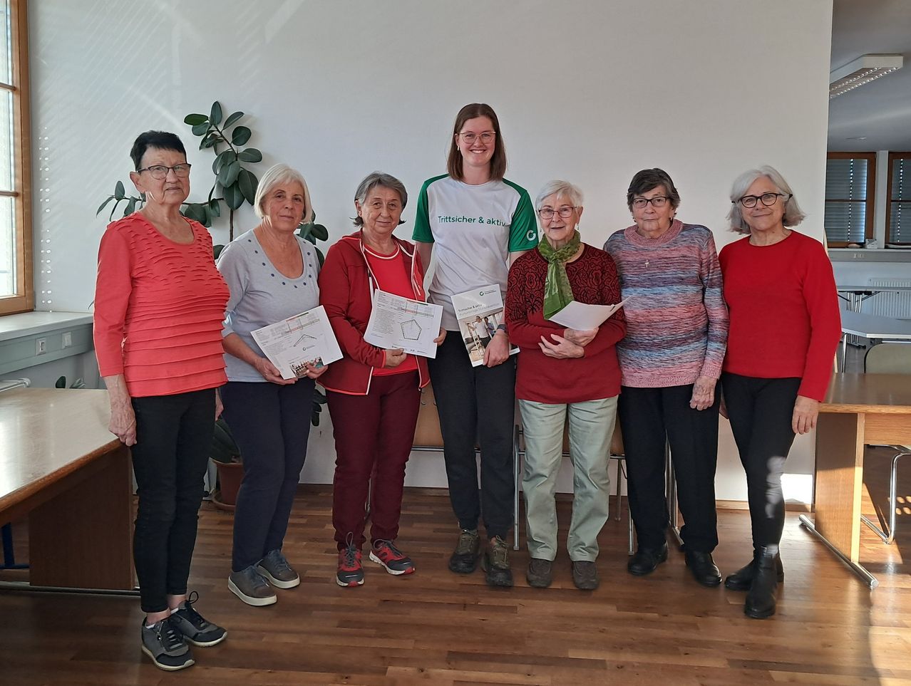 Sieben Frauen posieren für ein Foto in einem Raum, jede hält ein Papier in der Hand. Eine Frau in der Mitte trägt ein grün-weißes Shirt mit Text. Hinter ihnen befindet sich eine weiße Wand mit einer Topfpflanze.