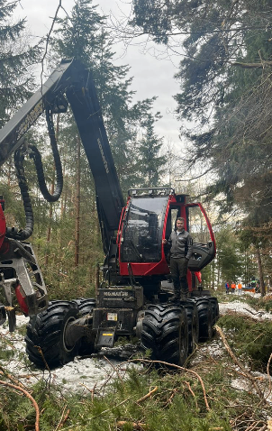 Ein Mann steht neben einer rot-schwarzen Forstmaschine in einem verschneiten Wald mit Bäumen im Hintergrund.