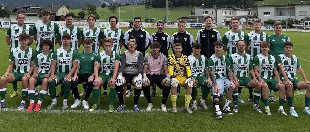 Eine Fußballmannschaft in grünen und weißen Trikots posiert für ein Gruppenfoto auf einem Rasenfeld mit Bergen im Hintergrund.