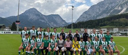 Eine Fußballmannschaft posiert für ein Gruppenfoto auf einem Feld mit Bergen im Hintergrund. Sie tragen grüne und weiße Trikots.