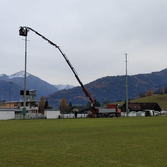 Ein Kran erstreckt sich über ein Feld mit Bergen im Hintergrund. Menschen gehen in der Nähe des Krans und Gebäude sind auf der linken Seite zu sehen.