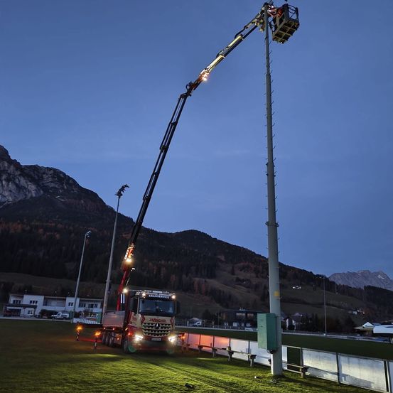 Ein Kranwagen steht auf einem Rasenfeld mit einem hohen Mast. Er ist beleuchtet, möglicherweise für Wartungsarbeiten. Im Hintergrund befinden sich Berge.