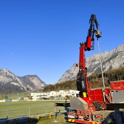 Ein großer roter Kran steht auf einem Feld mit Bergen im Hintergrund. Ein Sportplatz und mehrere Gebäude sind in der Ferne zu sehen.