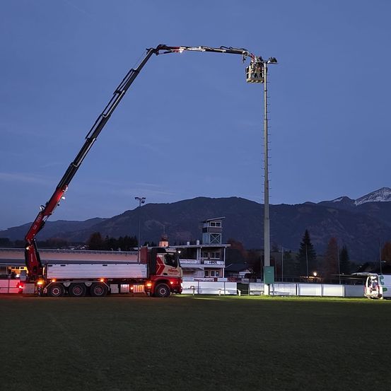 Ein großer Kran erstreckt sich auf einen hohen Mast zu, wobei ein Arbeiter den Kran bedient. Im Hintergrund sind eine Berglandschaft und ein Gebäude zu sehen.
