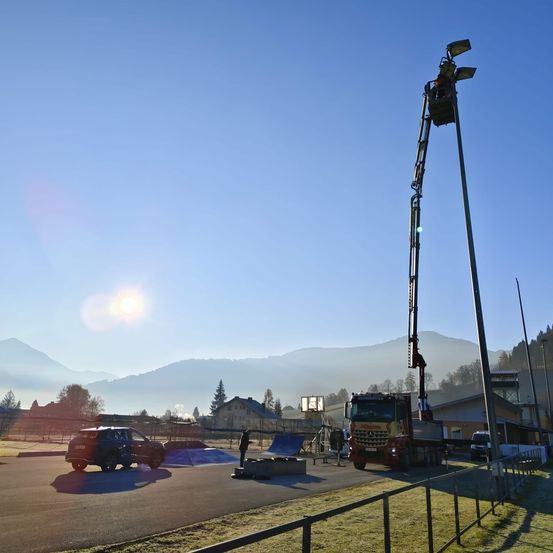 Ein sonniger Tag mit einem großen LKW, einer Person in einem Korblift und einem geparkten Auto in der Nähe eines Zauns und Gebäuden mit Bergen im Hintergrund.