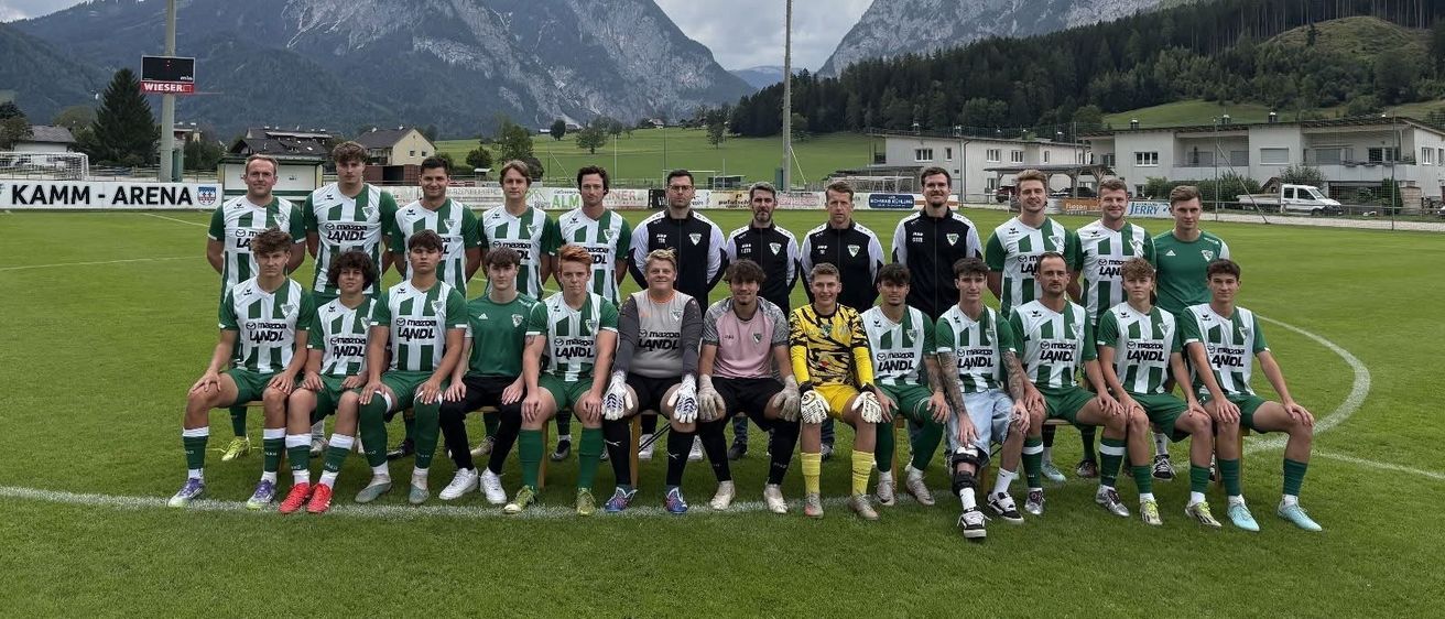 Eine Fußballmannschaft in grünen und weißen Trikots posiert für ein Gruppenfoto auf einem Feld mit Bergen im Hintergrund.