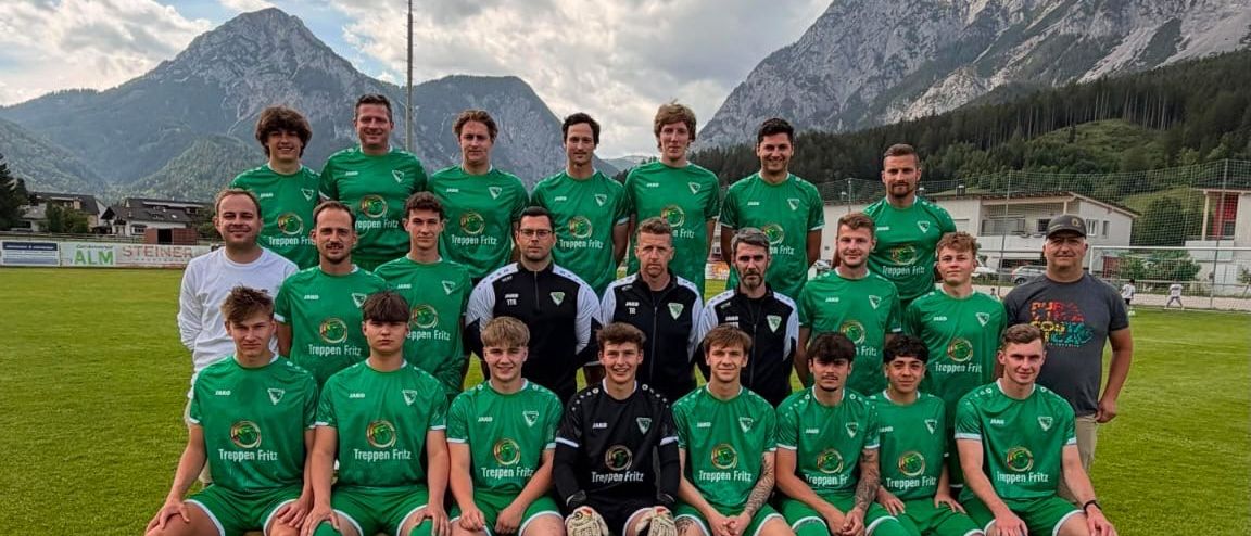 A soccer team in green uniforms poses for a photo on a field with mountains in the background.