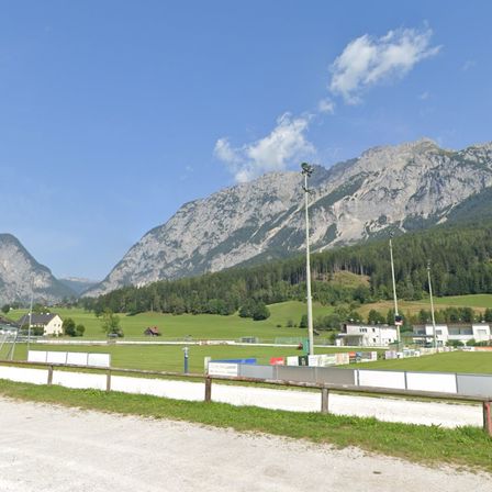 Ein Fußballfeld mit Zaun und Bergen im Hintergrund. Der Himmel ist blau mit einigen Wolken.