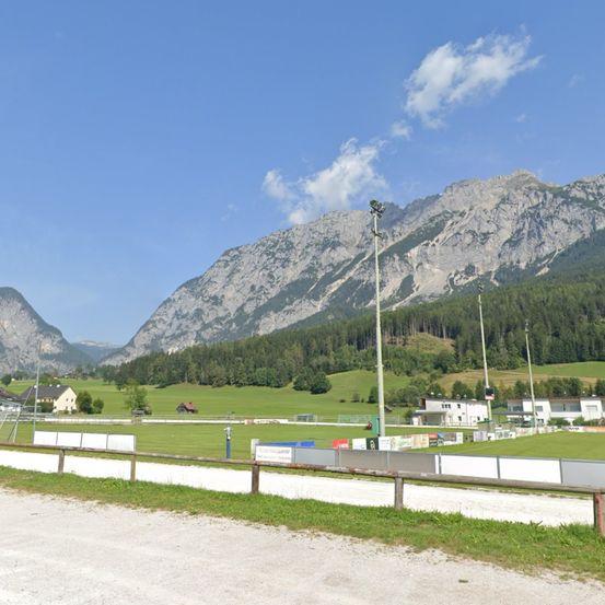 Ein Fußballfeld mit Zaun und Bergen im Hintergrund. Der Himmel ist blau mit einigen Wolken.