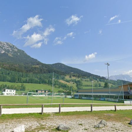 Ein Fußballfeld mit Netz und Stangen ist von einem Zaun umgeben. Hinter dem Zaun befindet sich ein Gebäude mit Dach und einem Berg in der Ferne.
