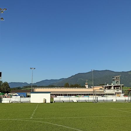 Ein großes Fußballfeld mit einem weißen Gebäude und Tribünen im Hintergrund, umgeben von Bergen und Straßenlaternen.