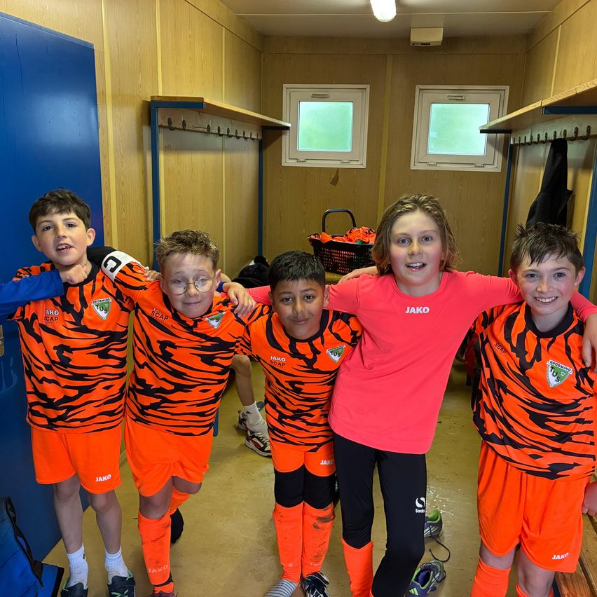 Fünf junge Fußballer, vier Jungen und ein Mädchen, stehen in einer Umkleidekabine und tragen orangefarbene Trikots. Das Mädchen hat die Arme um die Jungs gelegt, und sie alle sehen fröhlich aus.