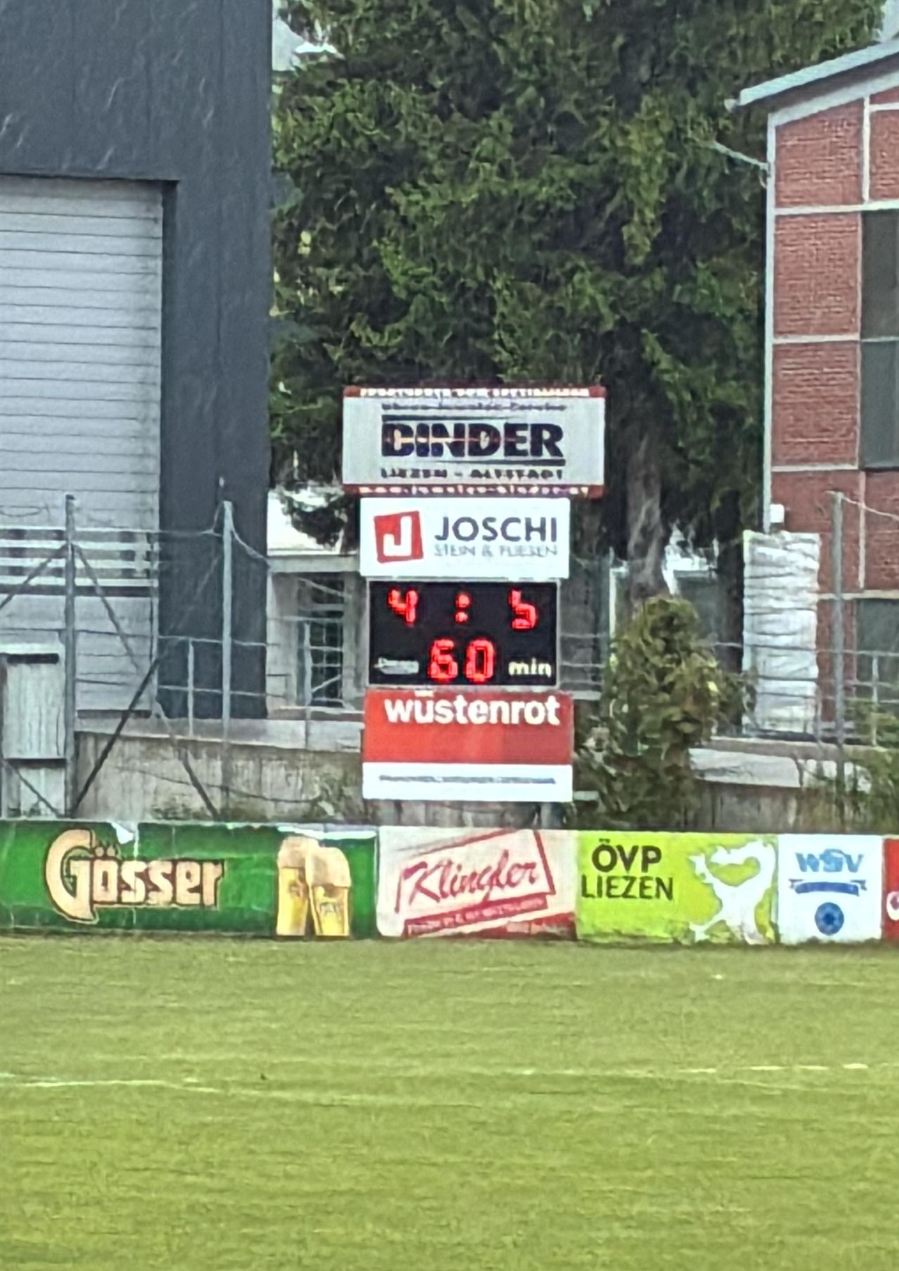 Ein Anzeigetafel mit der Anzeige von 4 zu 5 und 60 verbleibenden Minuten befindet sich auf einem Sportplatz. Im Hintergrund befinden sich Werbebanner, Bäume und Gebäude.
