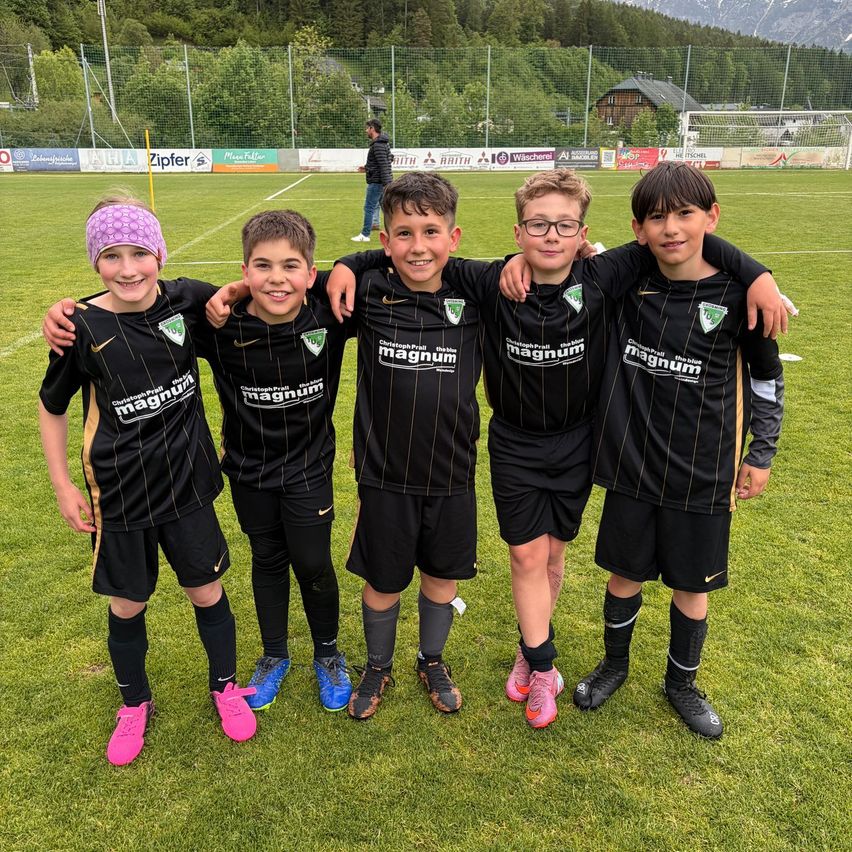 Fünf Jungen in Fußballtrikots stehen eng beieinander auf einem Feld. Sie lächeln und posieren für ein Foto. Dahinter steht ein Zaun mit einem Fußballtor.