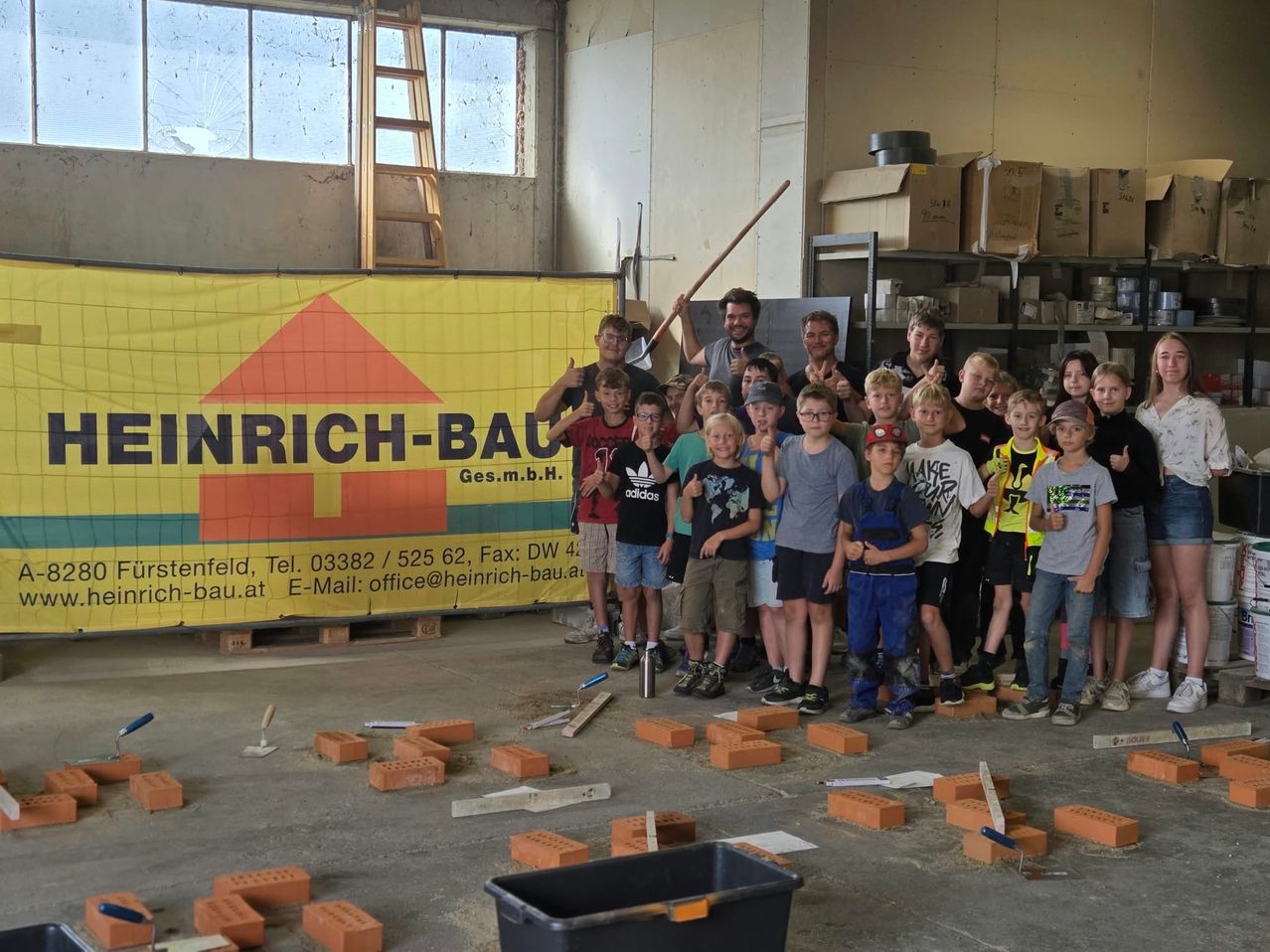 Eine Gruppe von Kindern und Erwachsenen steht in einem Raum mit Ziegeln und Werkzeugen und posiert für ein Foto vor einem Banner.