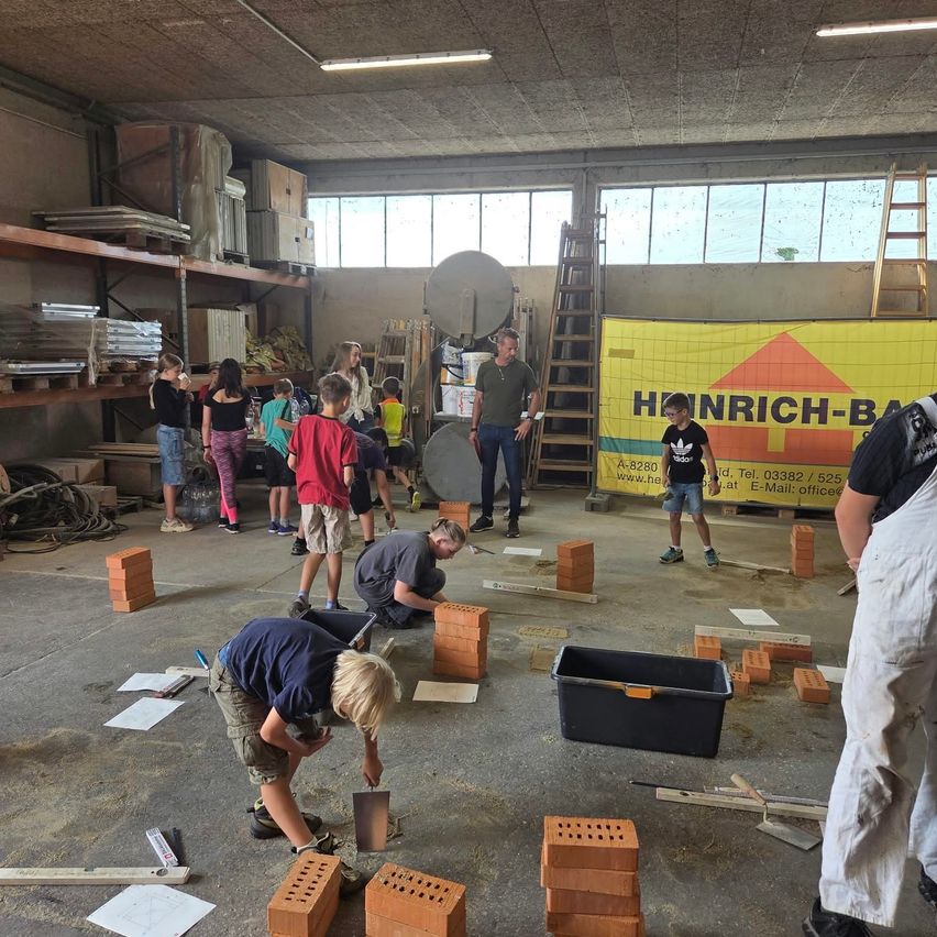 Eine Gruppe von Kindern und Erwachsenen arbeitet in einer Werkstatt an einem Ziegellegeprojekt. Einige knien auf dem Boden, während andere stehen und beobachten. An der Wand ist ein Banner mit dem Text 'HANRICH-BAU' angebracht.
