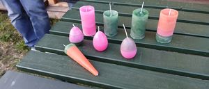 Auf einem Picknicktisch steht eine Reihe von Kerzen in verschiedenen Farben und Formen, darunter rosa, grün und orange. Eine Person steht hinter dem Tisch.